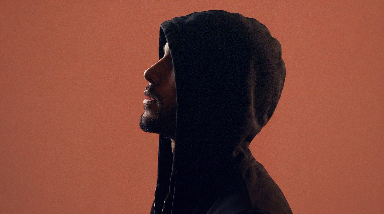 Profile portrait of Anik Devaughn in a black hood against a rust-orange background, looking up into the light.